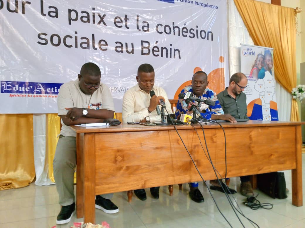 Quatre hommes sont assis derrière une table en bois devant une banderole sur la paix et la cohésion sociale au Bénin