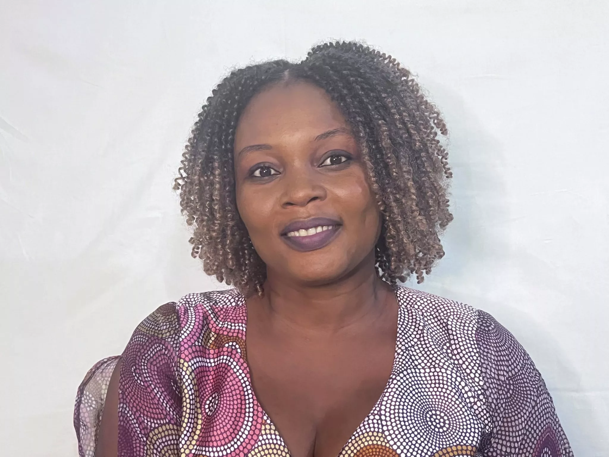 Femme souriante aux cheveux bouclés courts vêtue d'un haut coloré à motifs assise devant un fond blanc
