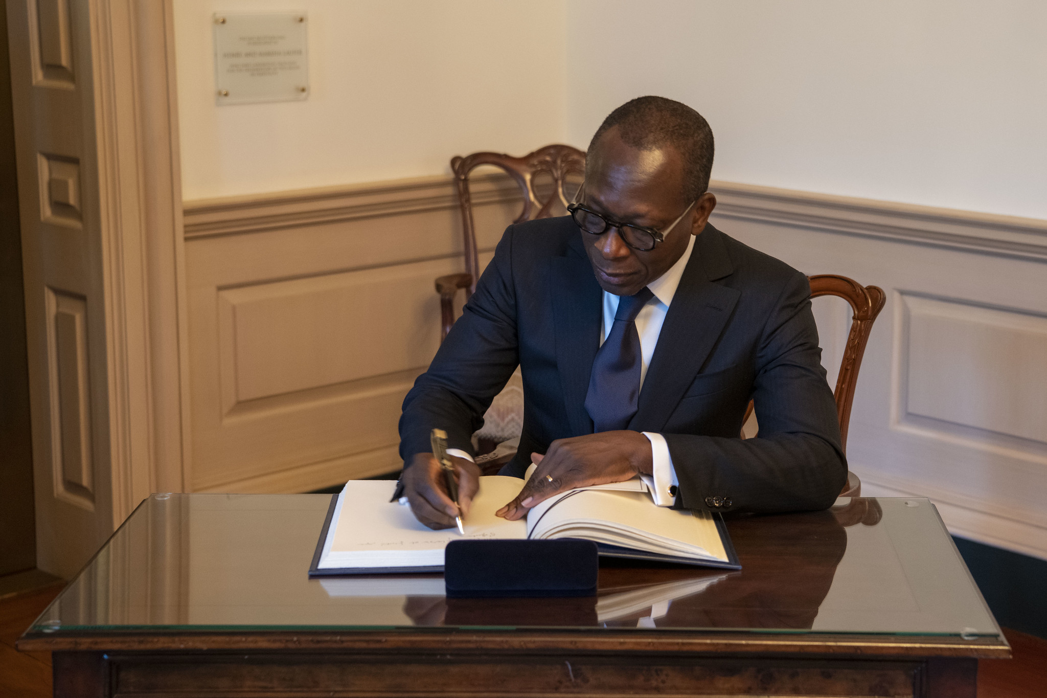 Un homme en costume sombre signe un document posé sur une table en bois dans une pièce à la décoration classique