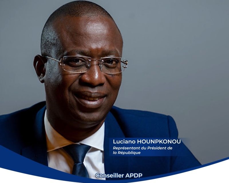 Homme en costume bleu et cravate souriant portant des lunettes sur fond gris avec texte descriptif en bas