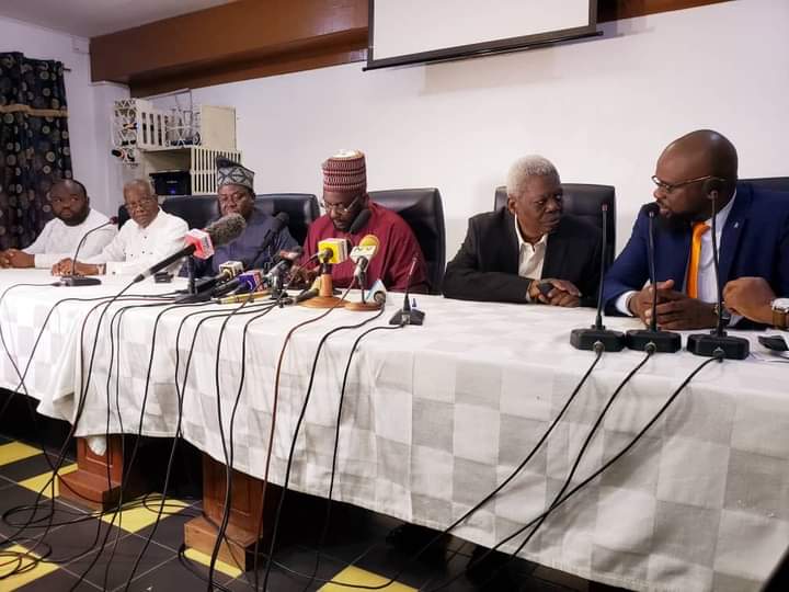 Six hommes assis à une table avec de nombreux microphones et appareils d'enregistrement devant eux dans une salle de réunion