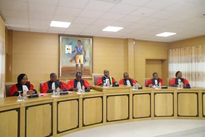Sept personnes en robe rouge et noire assises derrière une large table en bois dans une salle de réunion officielle