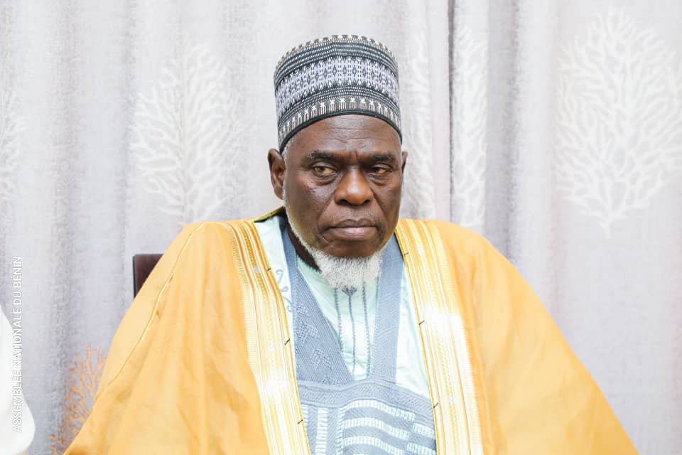 Homme portant une djellaba jaune et un bonnet traditionnel gris, assis devant un rideau clair à motifs discrets