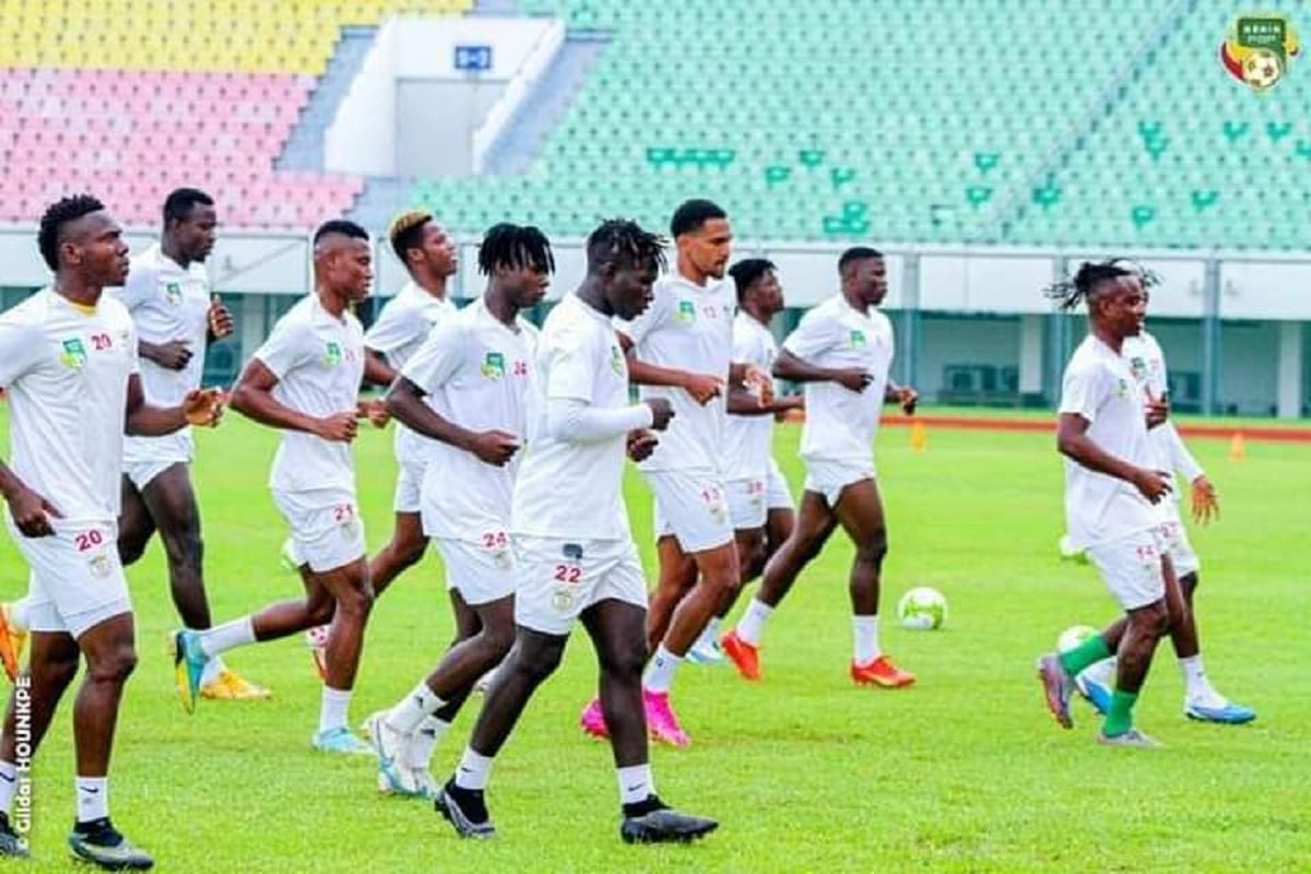 Des joueurs de football en tenue d’entraînement blanc courent sur une pelouse dans un stade aux gradins multicolores