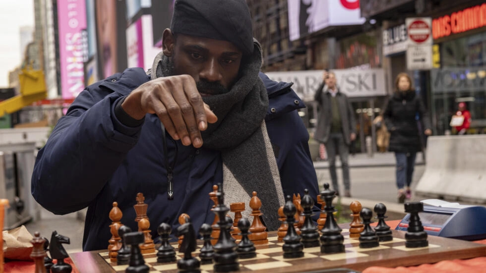 Un homme portant bonnet et écharpe joue aux échecs en pleine rue animée sur un échiquier en bois