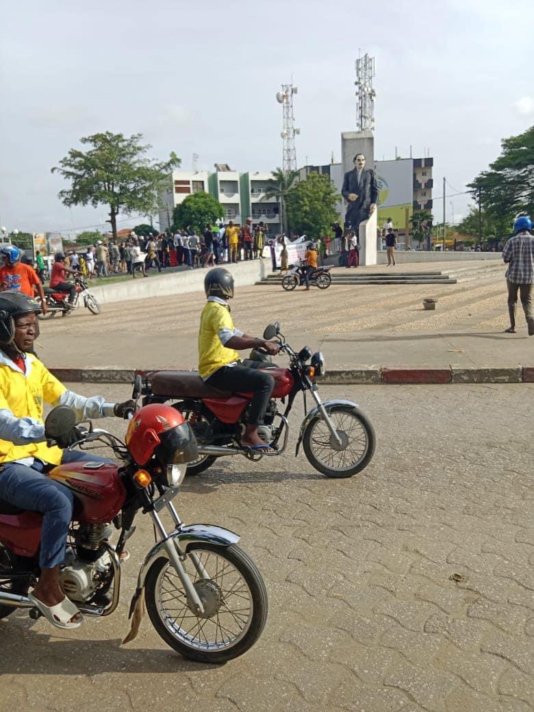 Deux hommes en moto passent devant une place avec une foule rassemblée près d'une statue imposante