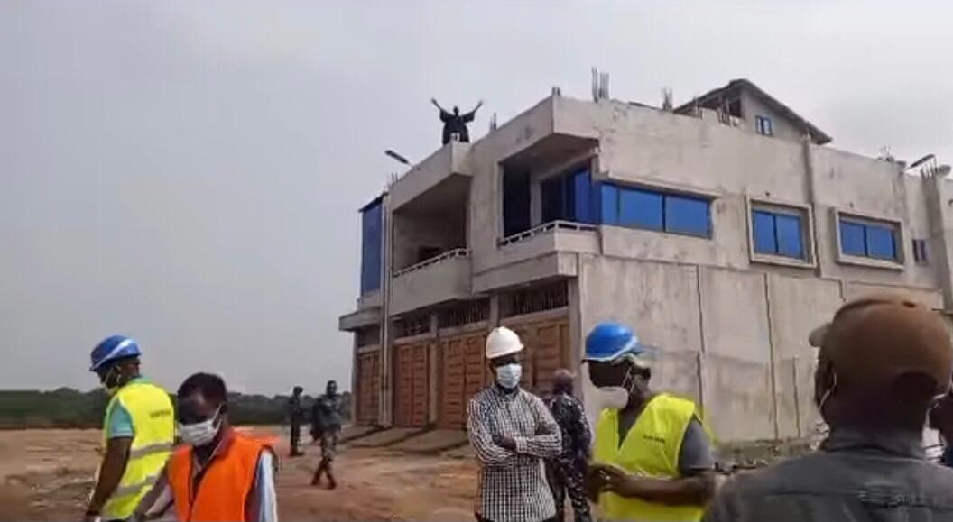 Des hommes portant des casques et des gilets fluorescents discutent devant un bâtiment inachevé avec une personne sur le toit levant les bras