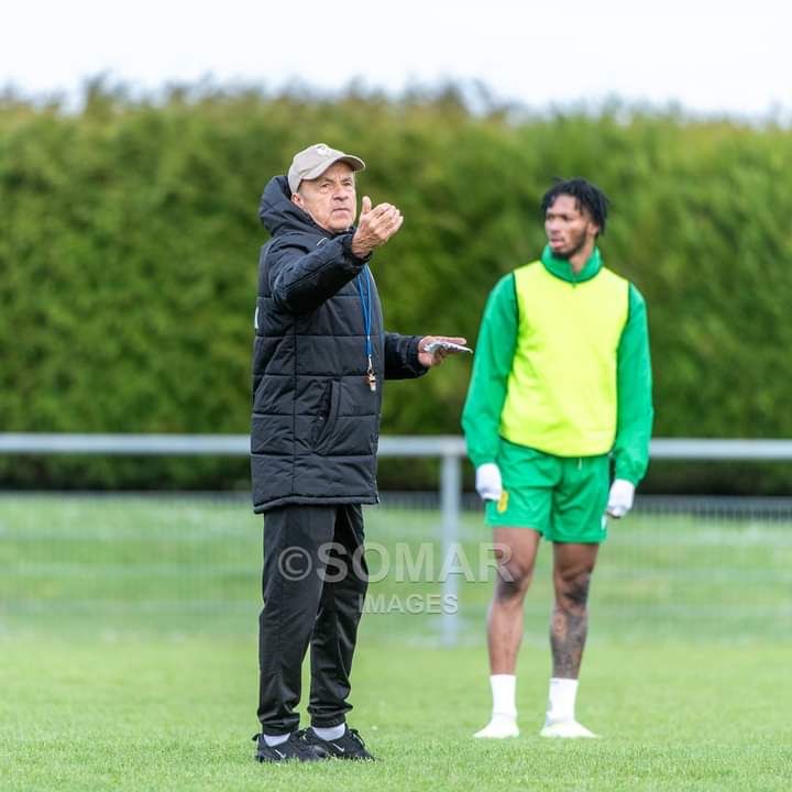 Un homme en tenue sportive noire et casquette donne des instructions à un joueur en tenue verte avec chasuble jaune sur un terrain herbeux