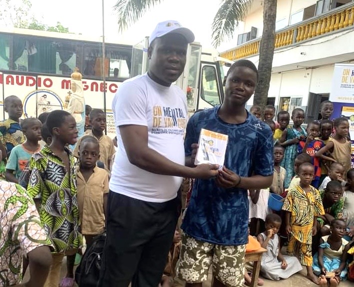Un homme en casquette blanche remet un livre à un adolescent devant un groupe d’enfants rassemblés à l’extérieur