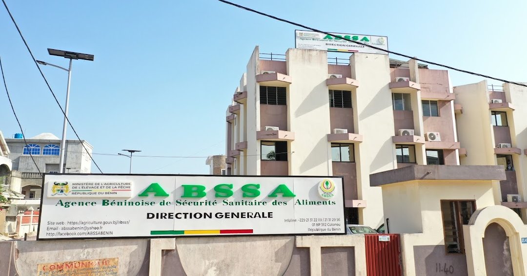 Bâtiment blanc de plusieurs étages avec panneau ABSSA Direction Générale devant un mur beige et un ciel bleu clair.