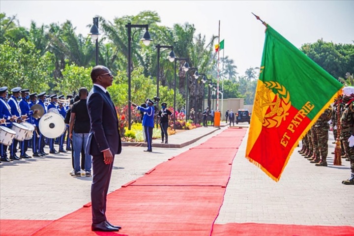 Un homme debout face à un drapeau, militaires en tenue et musiciens alignés de chaque côté d’un tapis rouge en extérieur