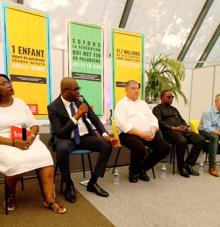 Cinq personnes assises en conférence dont un homme tenant un micro avec trois affiches sur la lutte contre le paludisme en arrière-plan