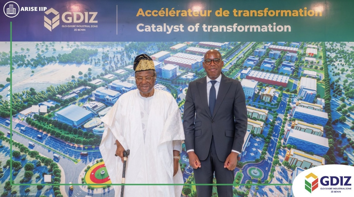 Deux hommes debout, l’un en tenue traditionnelle blanche et l’autre en costume sombre, devant une grande image aérienne colorée d’une zone industrielle