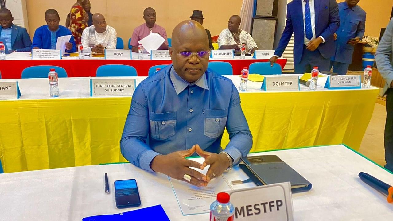 Un homme en chemise bleue assis à une table avec plusieurs documents et une bouteille d'eau lors d'une réunion officielle