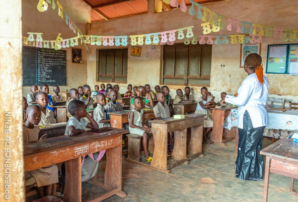Une enseignante debout face à une classe attentive d'enfants assis sur des bancs en bois dans une salle décorée de guirlandes en papier