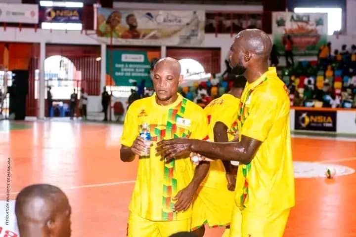 Trois joueurs en tenue jaune discutent et boivent sur un terrain de sport couvert avec tribune en arrière-plan