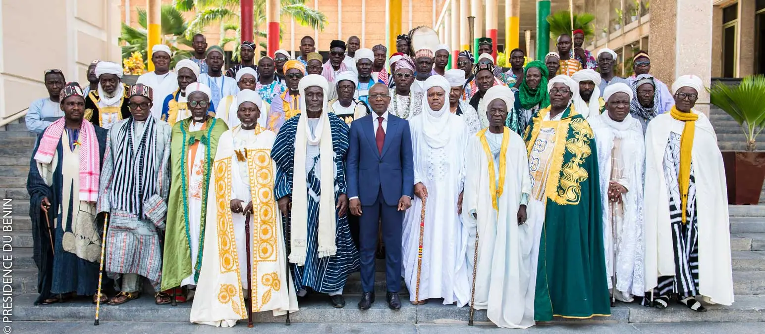 Groupe d’hommes en tenue traditionnelle et un homme en costume posant sur des marches devant un bâtiment avec des piliers colorés