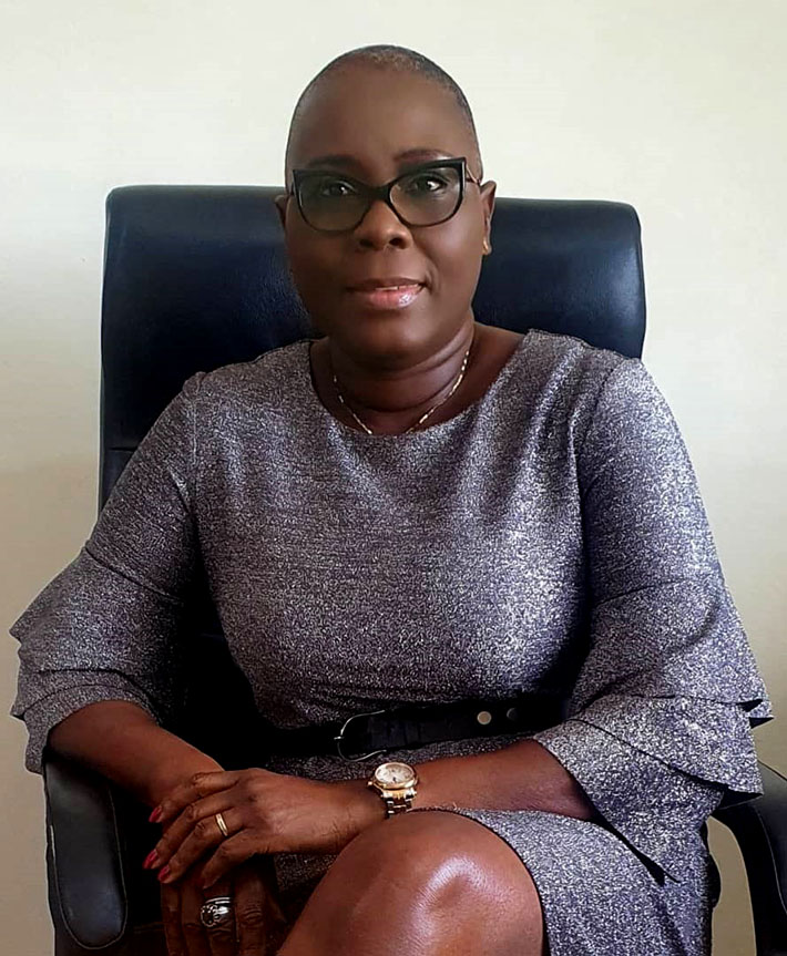 Femme assise en tailleur dans un fauteuil, portant des lunettes et une robe grise à manches évasées