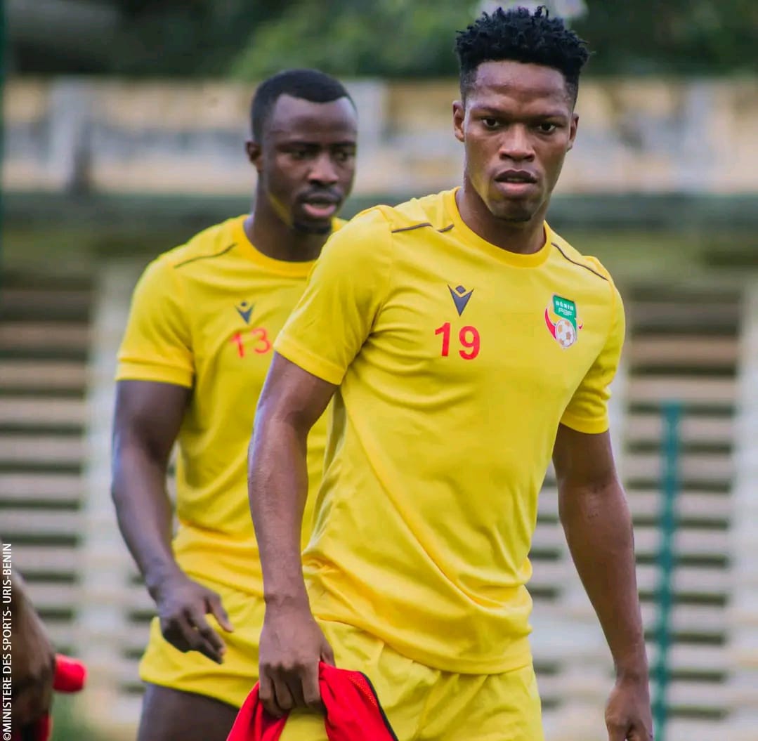 Deux joueurs de football en tenue d'entraînement jaune avec numéro rouge sur une pelouse devant des gradins vides