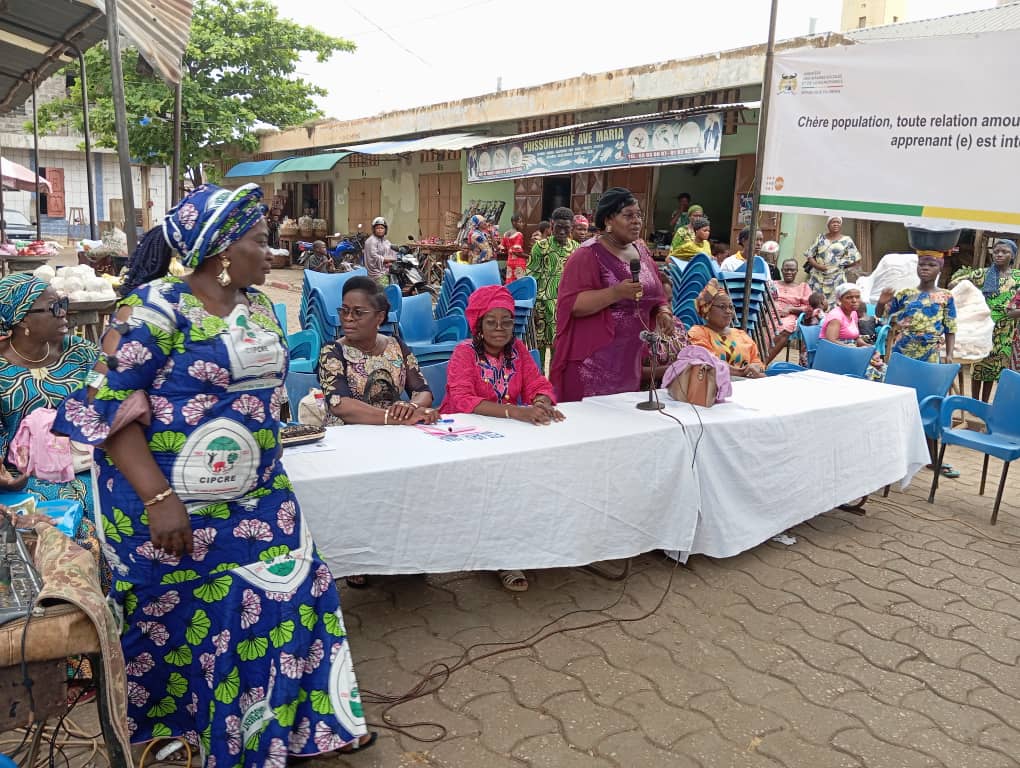 Une femme parle au micro derrière une table, entourée d’autres femmes assises et d’un public dans une rue piétonne en plein jour