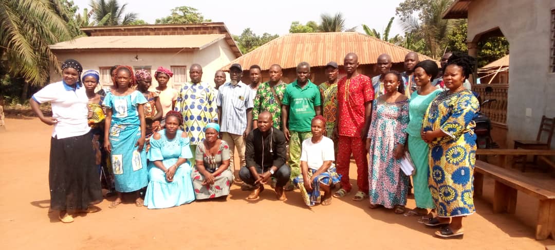 Groupe d’hommes et de femmes posant devant des maisons traditionnelles en milieu rural sous un ciel clair