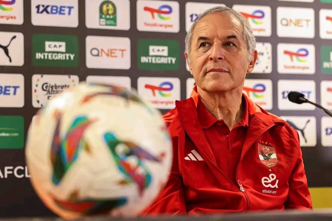 Homme en veste sportive rouge assis devant un micro avec un ballon de football flou au premier plan et logos sur fond noir