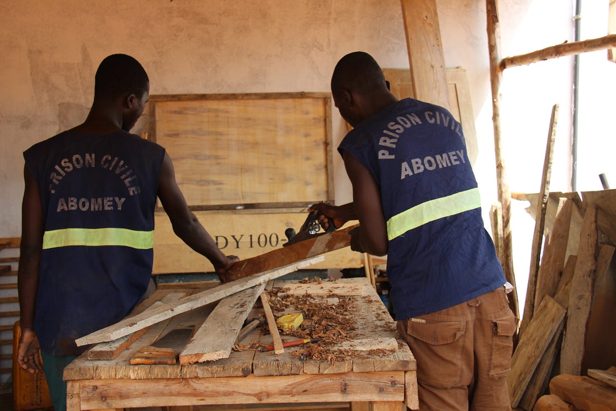 Deux hommes en gilet "Prison civile Abomey" travaillent le bois dans un atelier éclairé par une fenêtre à droite