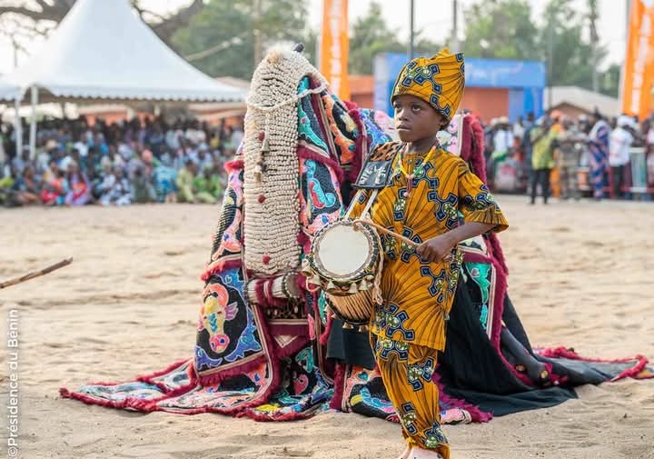 Enfant vêtu de pagne coloré jouant d’un tambour lors d’une cérémonie au sol sablonneux avec un personnage costumé en arrière-plan.