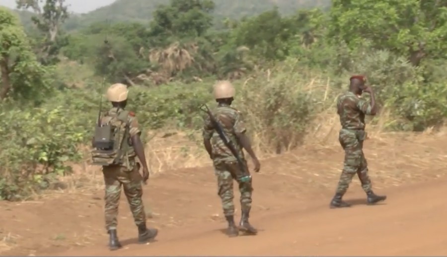 Trois soldats en tenue de camouflage et casque marchent sur une piste en terre entourée de végétation dense