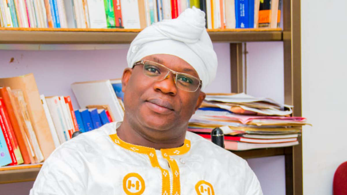 Homme portant un turban blanc et une tenue traditionnelle blanche brodée, assis devant une bibliothèque remplie de livres.