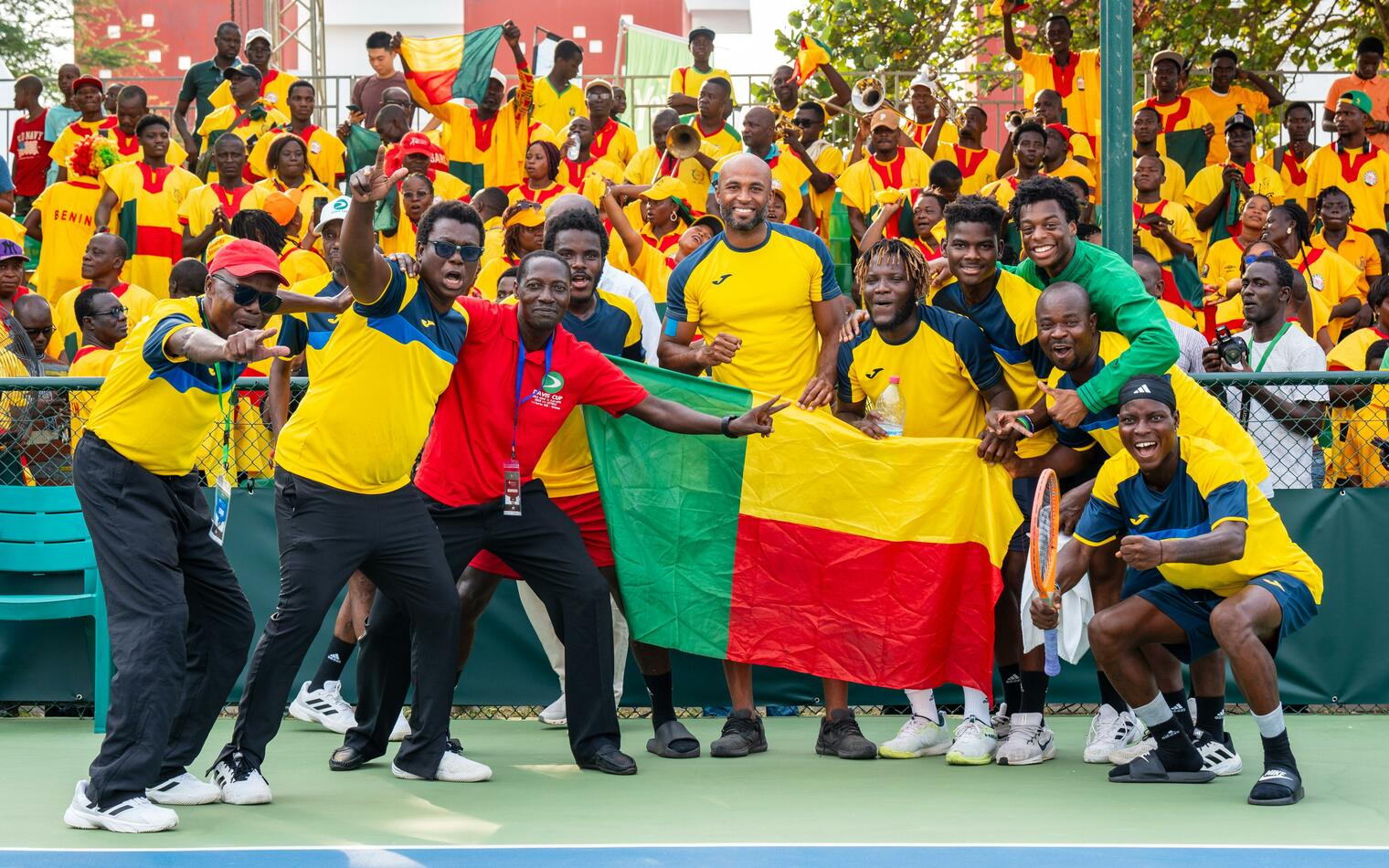 Un groupe d’hommes en tenue sportive jaune et verte brandit un drapeau tricolore rouge, jaune et vert sur un terrain de tennis avec un public en fond vêtu de ja