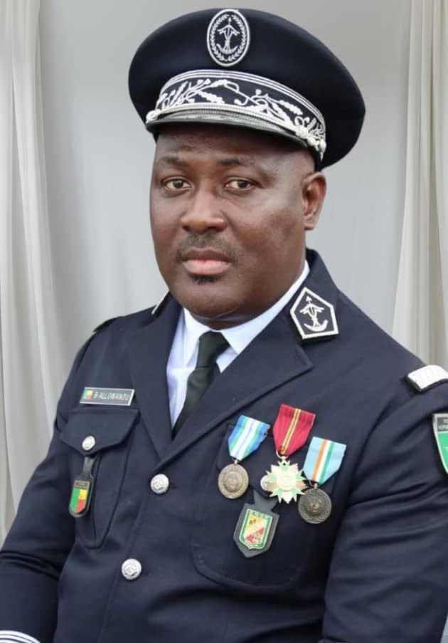 Homme en uniforme officiel orné de médailles et insignes posant devant un rideau clair
