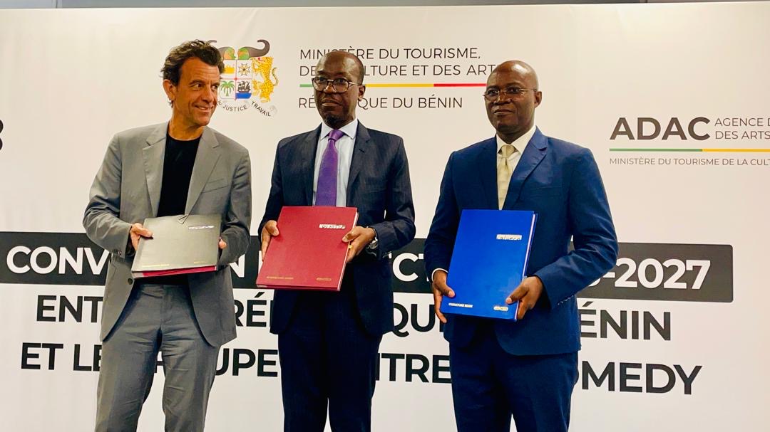 Trois hommes en costume tenant des dossiers colorés posent devant un mur portant des logos et du texte officiel