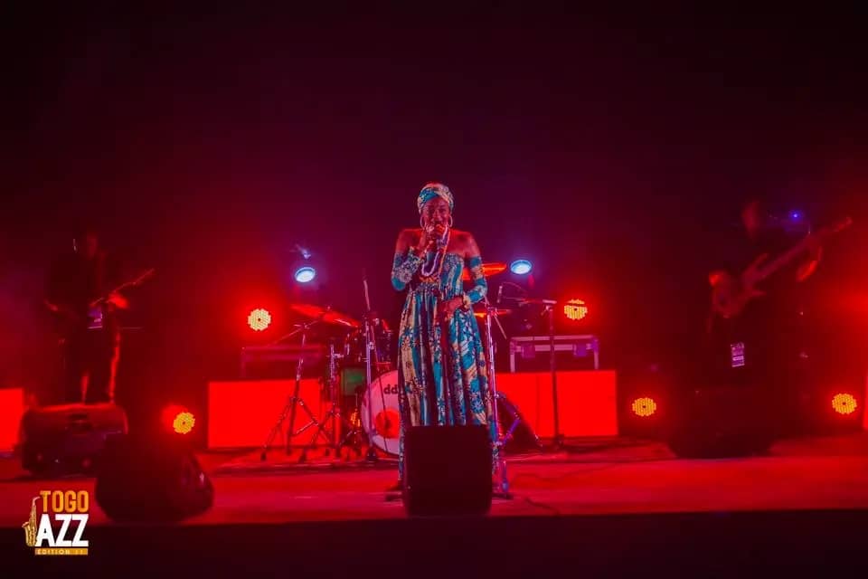 Chanteuse en tenue traditionnelle colorée sur scène éclairée en rouge avec musiciens en arrière-plan et logo Togo Jazz visible