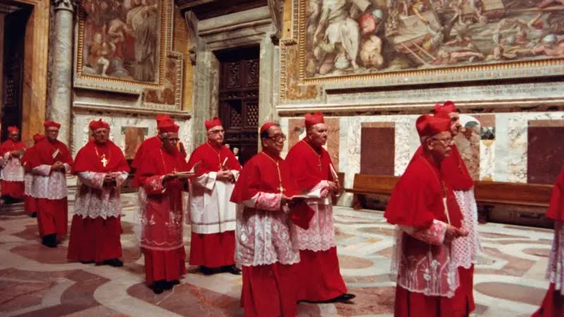 Un groupe de religieux en robe rouge avance en procession dans une grande pièce ornée de fresques murales.