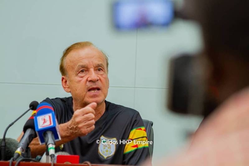 Un homme en maillot de sport noir parle en gesticulant devant un micro lors d’une conférence de presse