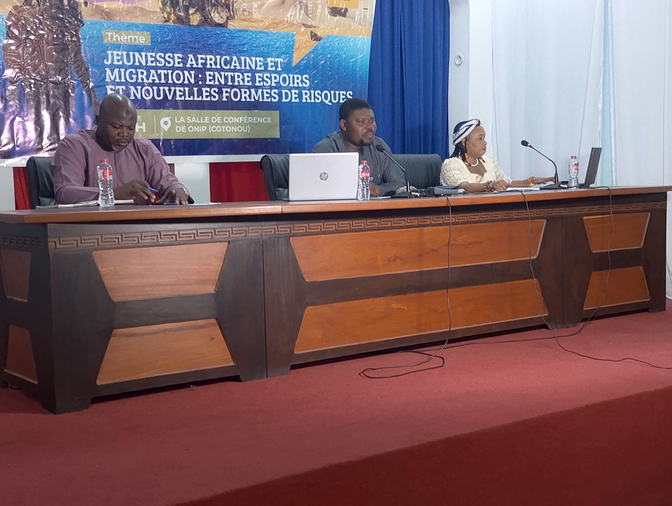 Trois personnes assises derrière une table de conférence avec un panneau thématique sur la jeunesse africaine et la migration en arrière-plan