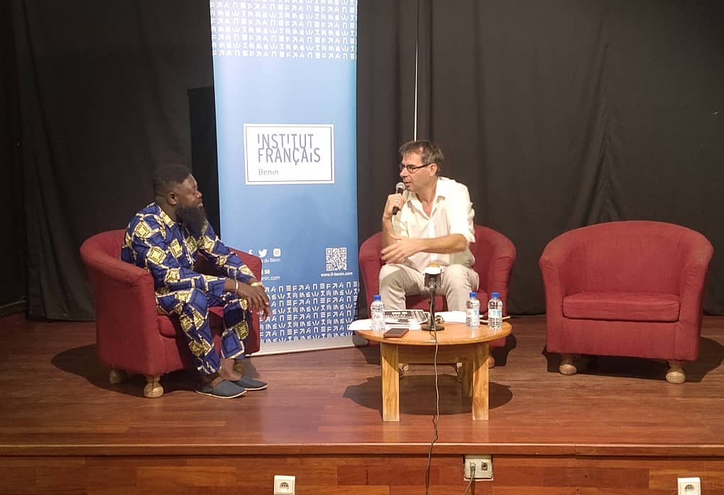 Deux hommes assis sur des fauteuils rouges sur une scène discutent face à un roll-up bleu de l'Institut Français et une petite table en bois devant eux