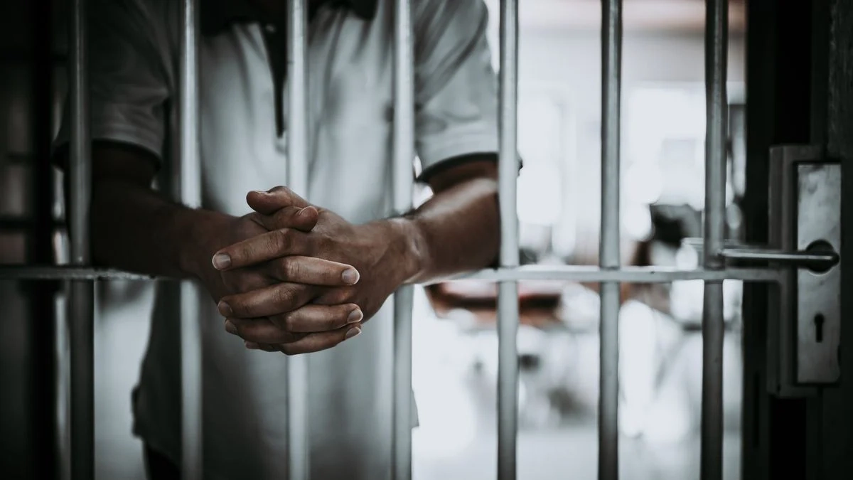 Mains croisées d’un homme en tenue claire agrippant les barreaux d’une cellule de prison avec porte fermée visible à droite