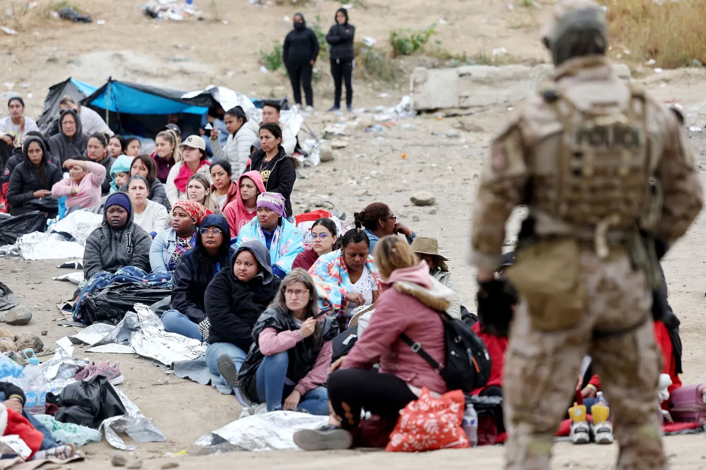 Un groupe de migrants assis sur le sol recouvert de couvertures de survie sous la surveillance d’un agent en tenue militaire