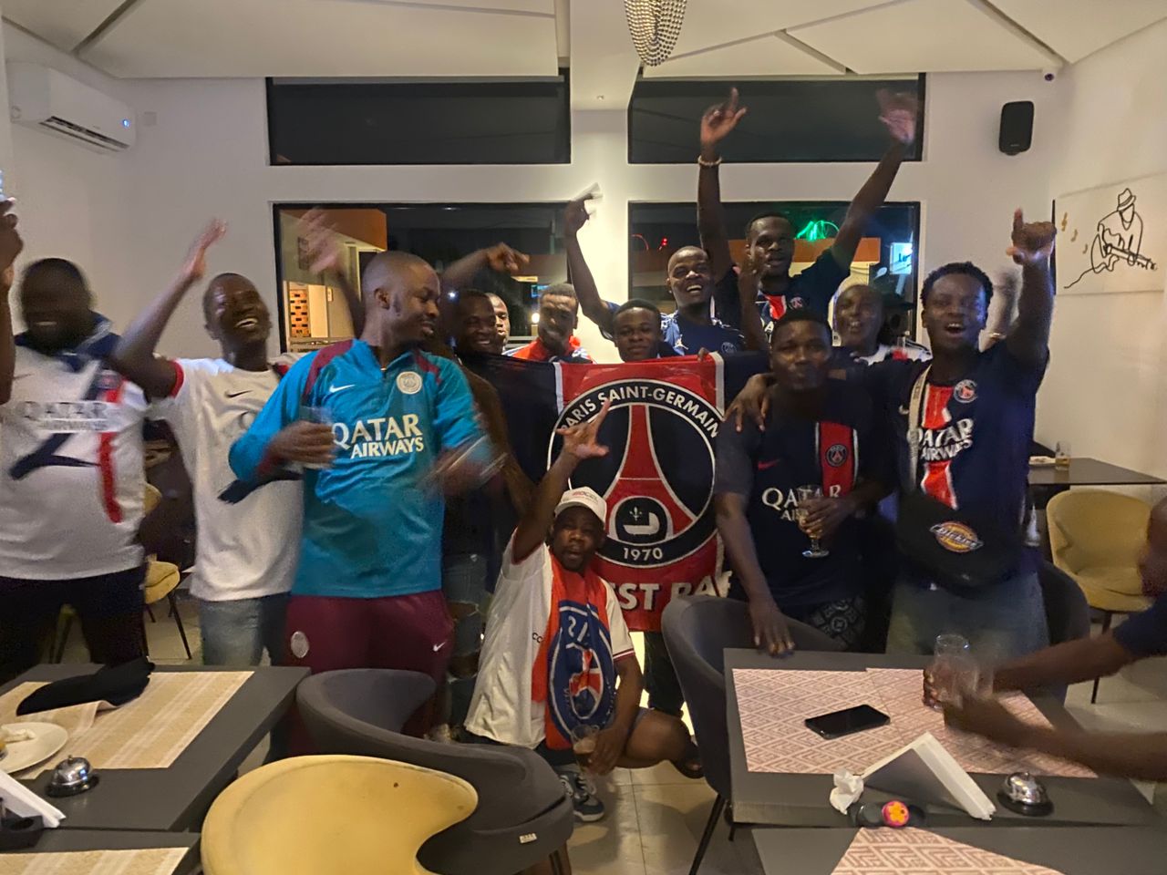 Groupe d'hommes célébrant avec un drapeau Paris Saint-Germain dans une salle avec tables et chaises