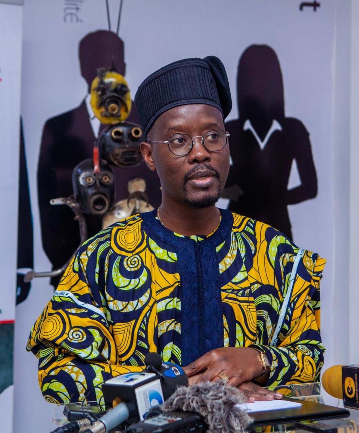 Homme en tenue traditionnelle colorée parlant devant plusieurs micros face à un fond avec silhouettes et objets d'art africain