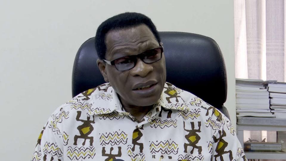 Un homme portant des lunettes et une chemise à motifs est assis dans un fauteuil devant une étagère avec des livres empilés