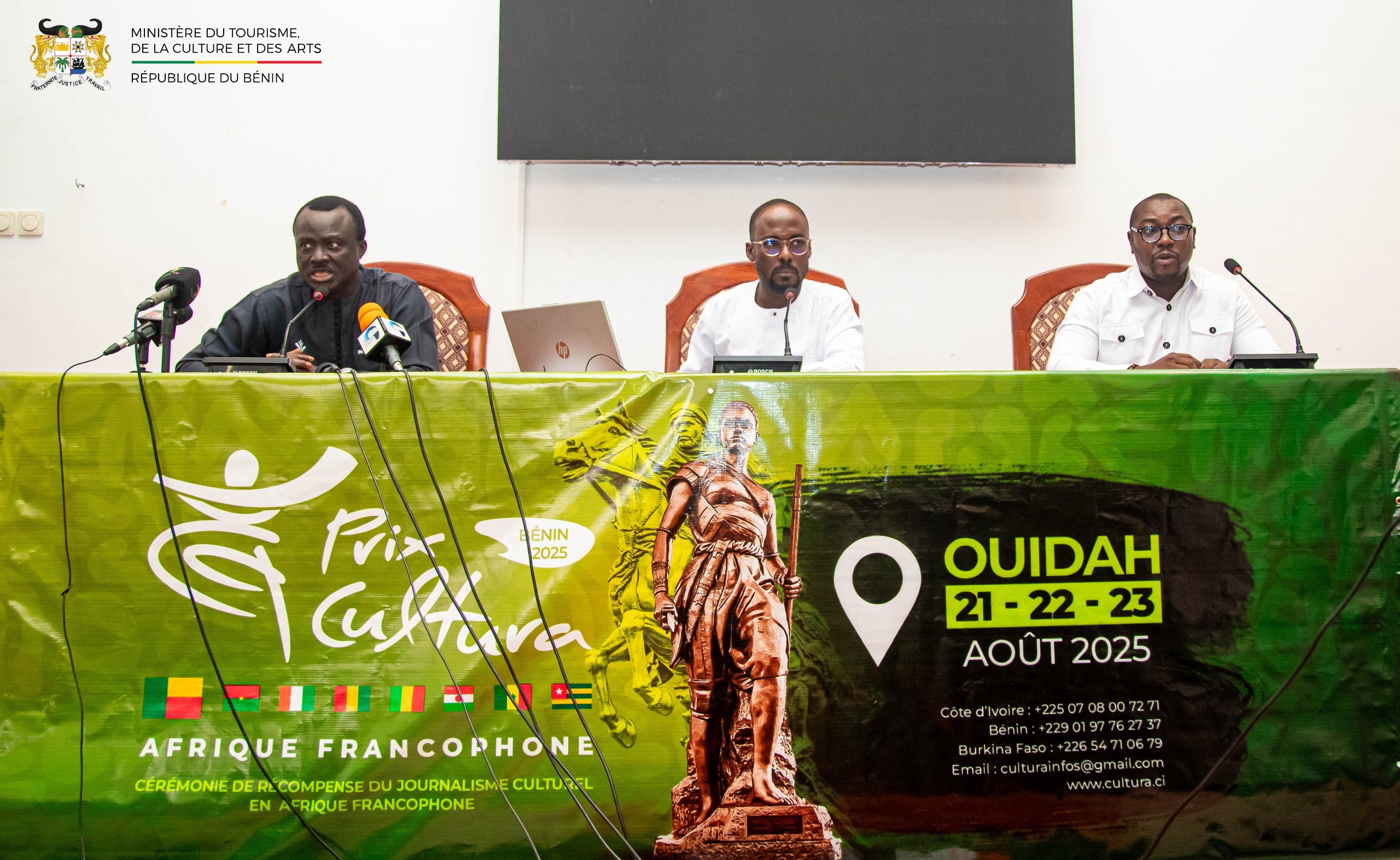 Trois hommes assis derrière une table verte avec micro, devant une bannière annonçant un événement en août 2025 à Ouidah