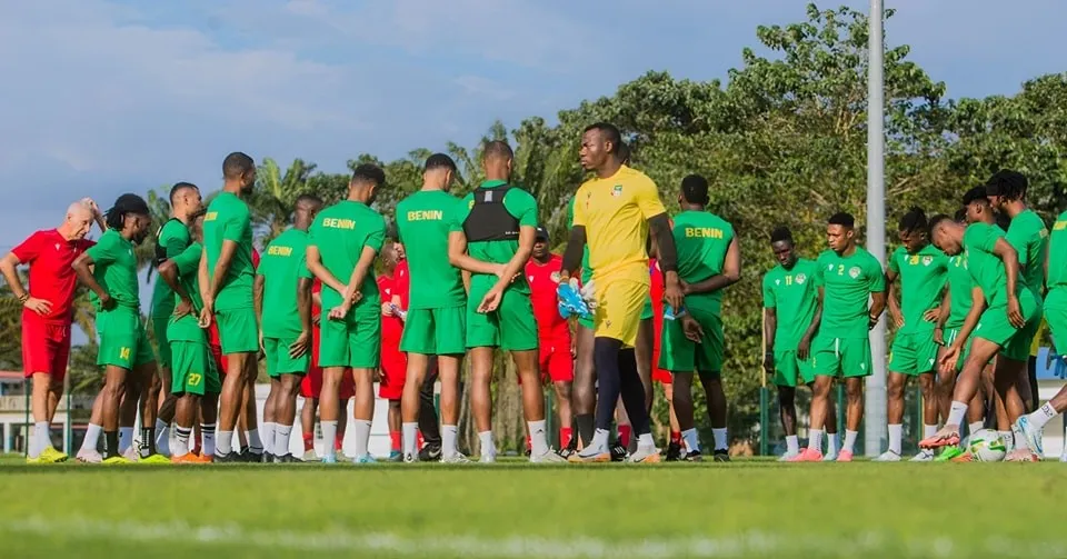 Des joueurs en tenue verte et quelques encadreurs discutent sur un terrain de football sous un ciel bleu.