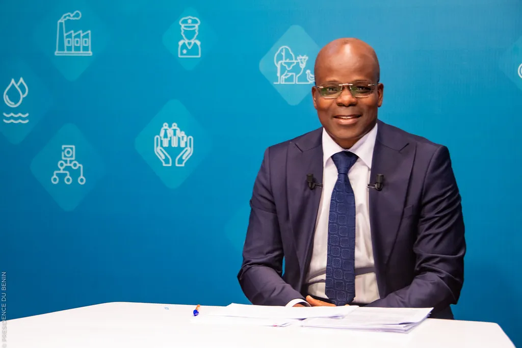 Homme en costume sombre et cravate souriant assis face à une table blanche sur fond bleu avec pictogrammes blancs