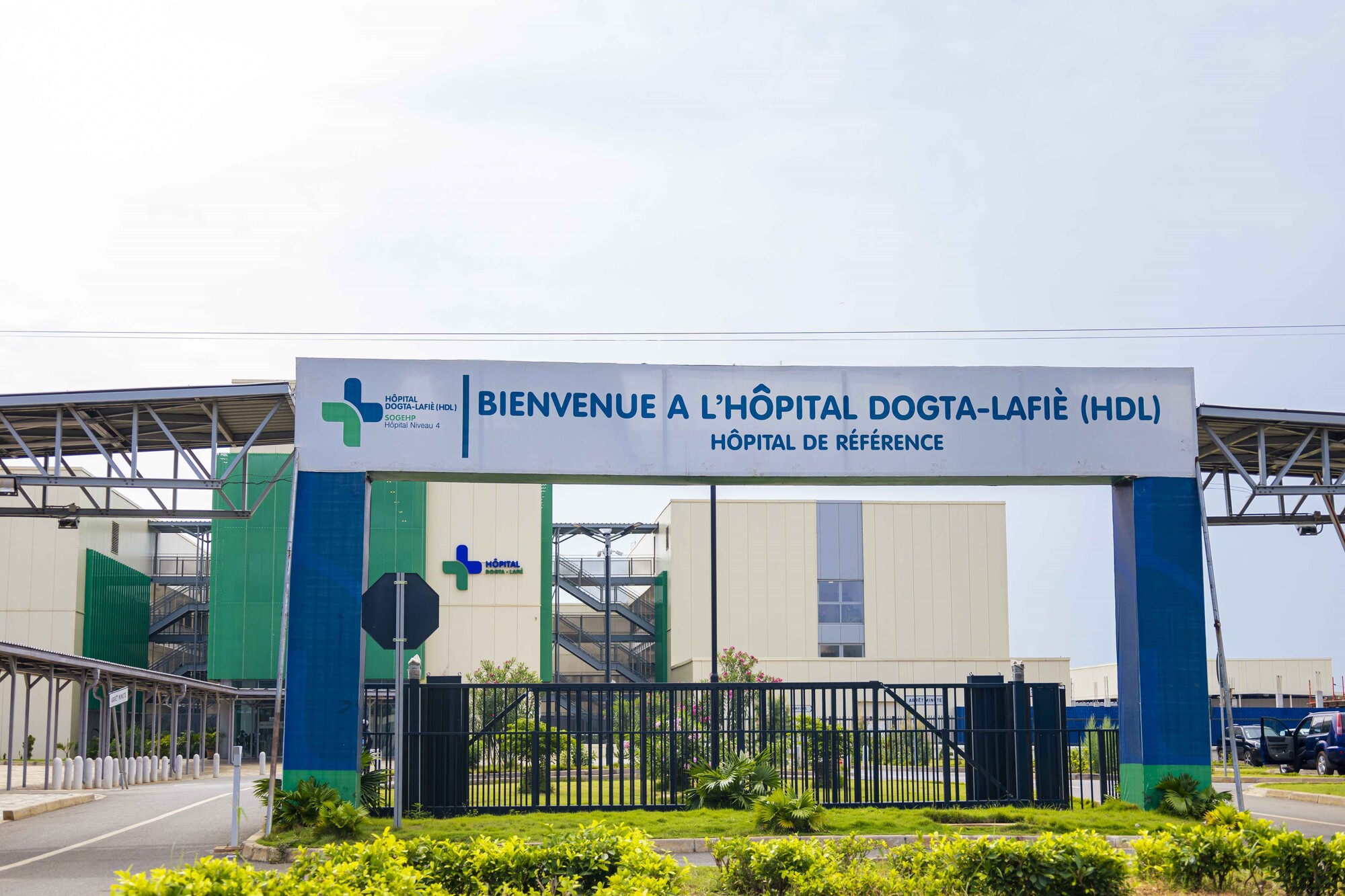 Entrée principale d’un hôpital moderne avec un portail, une barrière fermée et des plantes au premier plan