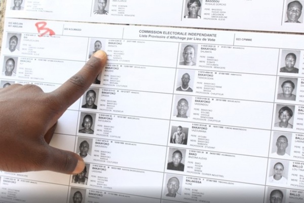 Main masculine pointant une ligne sur une liste électorale affichée avec photos et informations des électeurs