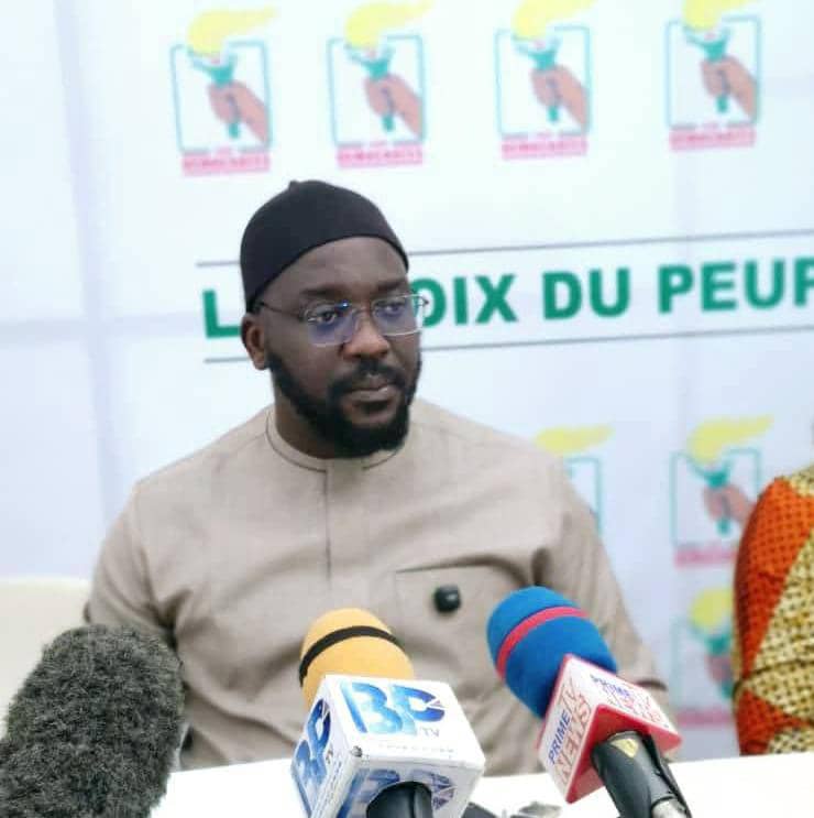 Homme portant un vêtement clair et un bonnet noir assis devant plusieurs micros et un fond avec des logos et un texte partiel "LA VOIX DU PEUPL