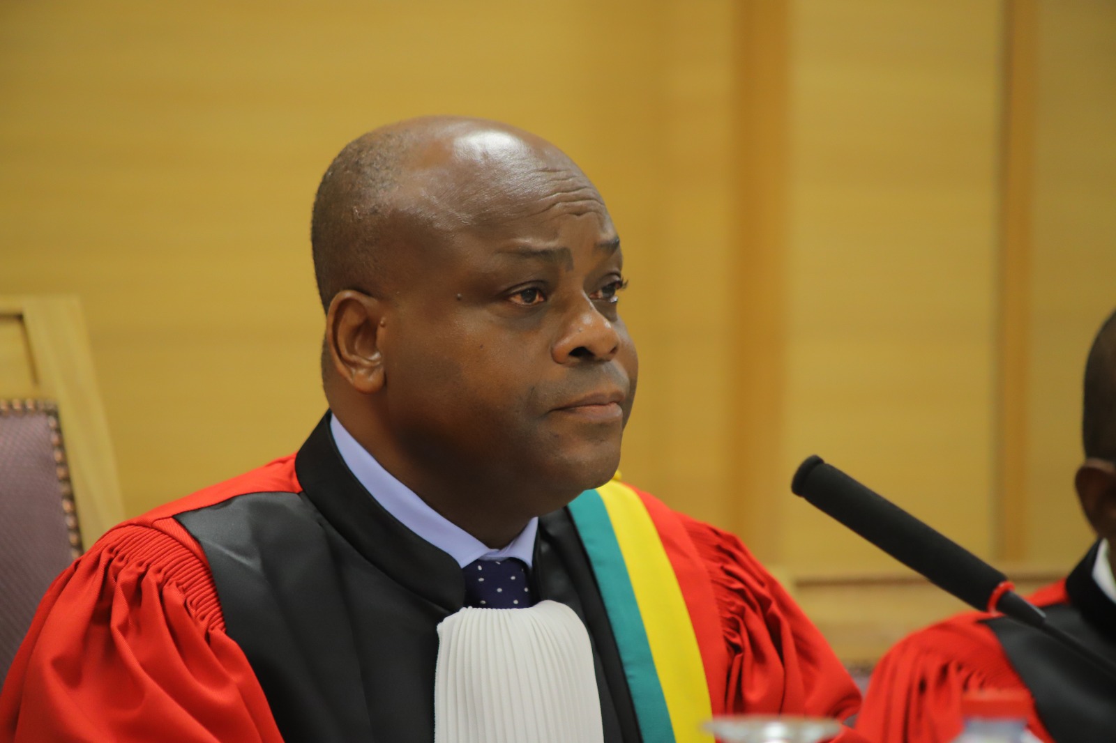 Homme en toge judiciaire rouge portant une écharpe tricolore dans une salle d’audience devant un micro